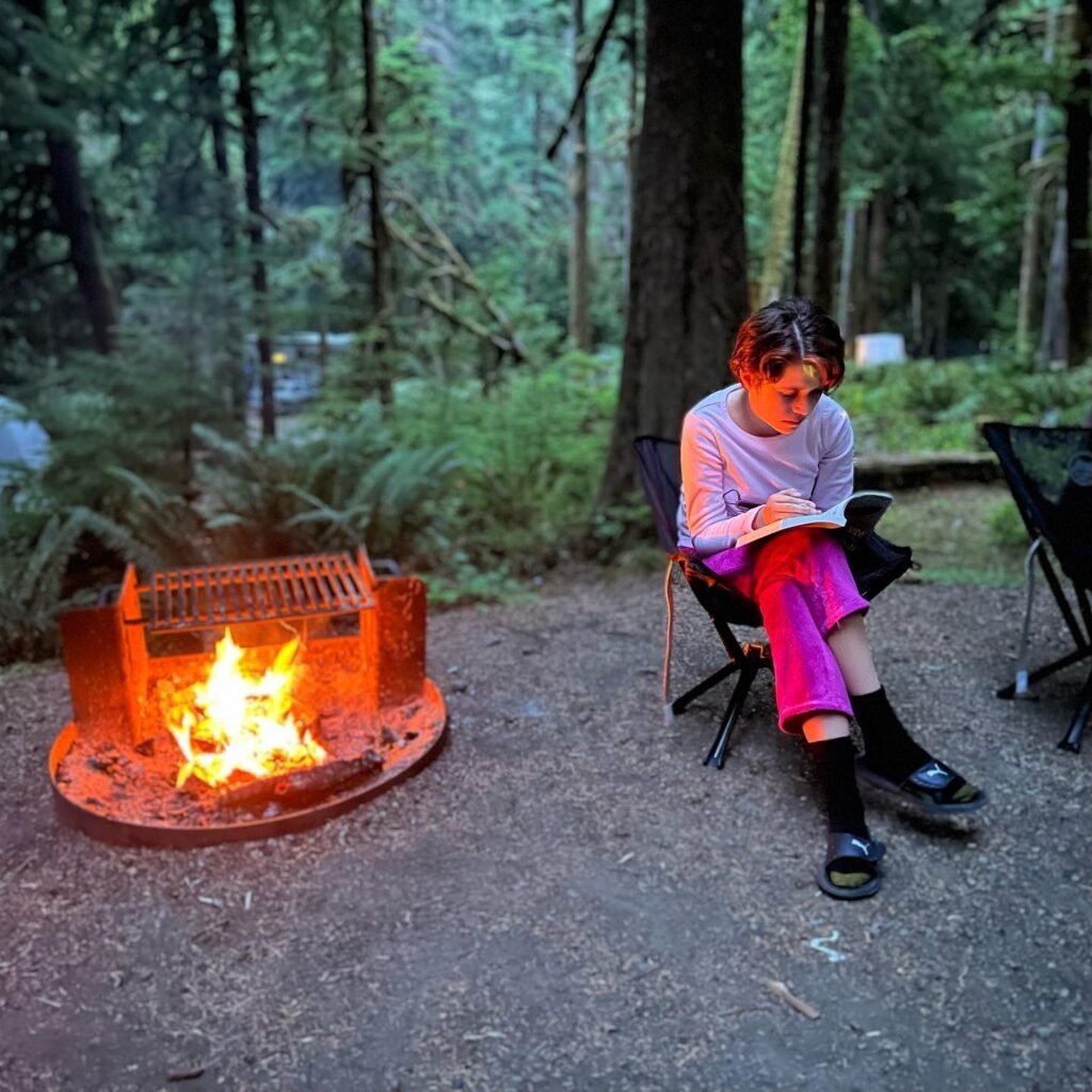Person sitting in a camp chair by a burning fire pit, reading a book in a forest setting.