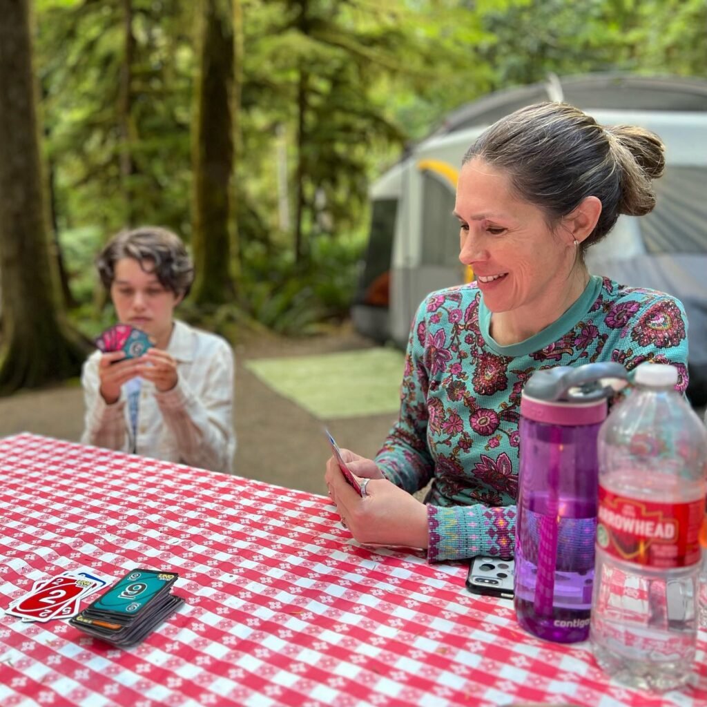 A woman and a young person playing card games at a red and white checkered picnic table in a forest campsite.