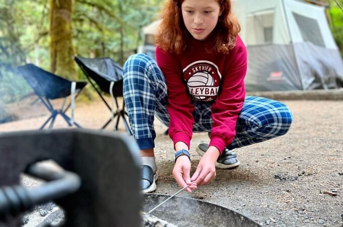 A young girl skillfully making s'mores by a campfire at a family campsite, surrounded by forested nature.