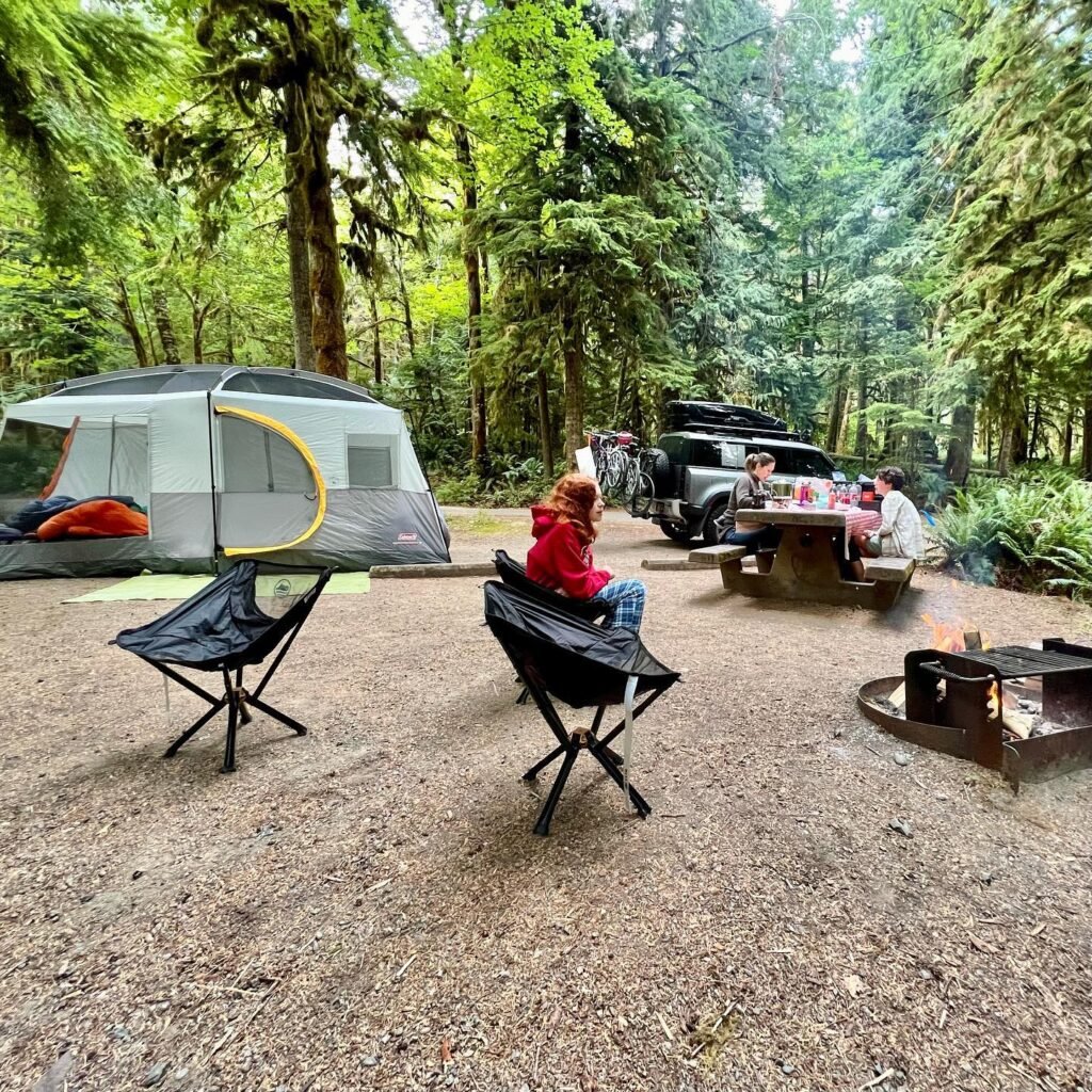 A cozy campsite in a lush Pacific Northwest forest with a tent, fire pit, and vehicle.