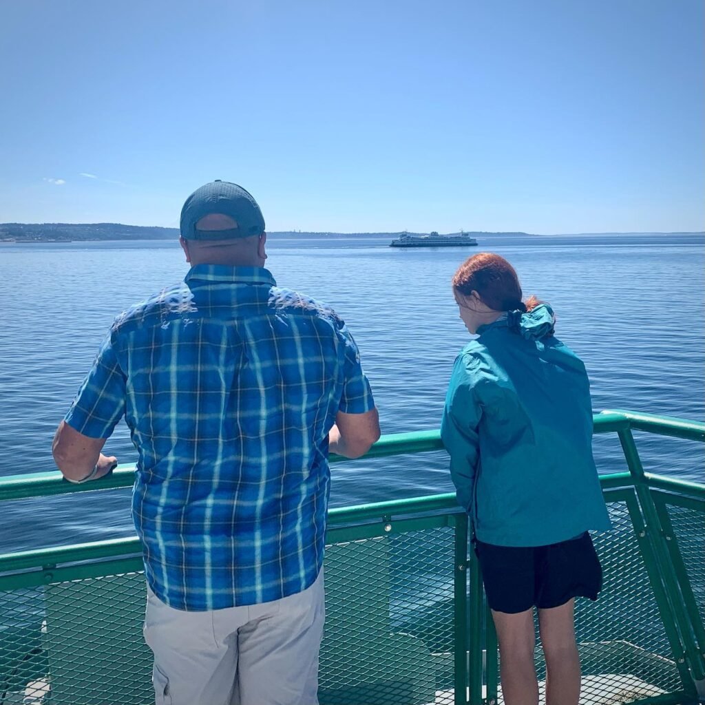 Two people stand at the railing of a ferry, gazing over calm blue waters toward a distant shoreline under a clear sky.