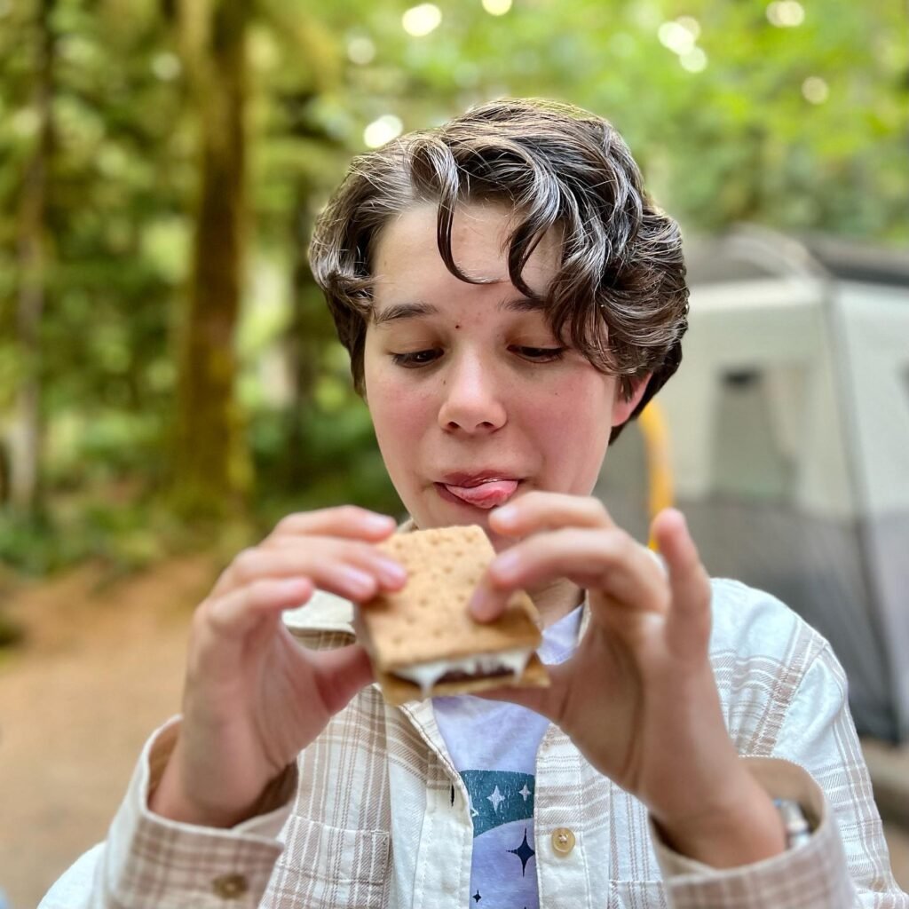 A person holds a sandwich with a wooded area and travel trailer in the background.