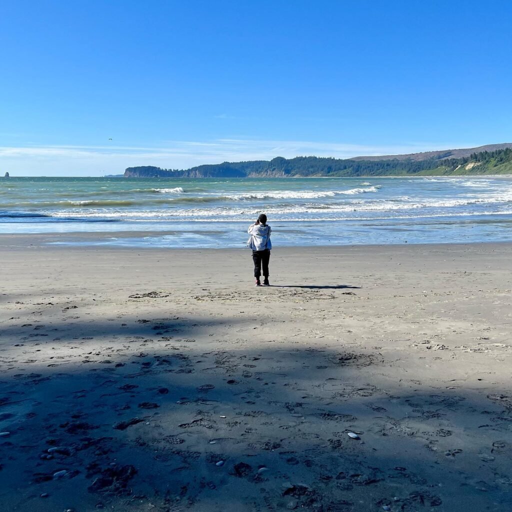 A person stands on a wide sandy beach facing the ocean. Rolling waves meet the shore under a bright blue sky.