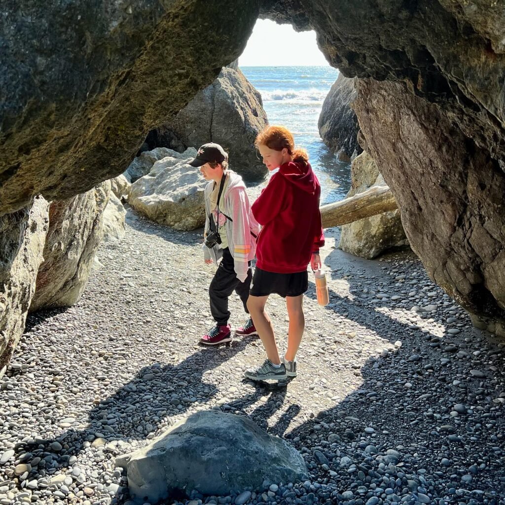 Hikers walk through a rocky cave passage opening into the ocean. Two people are near a stone archway overlooking the sea.