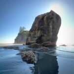 Sol Duc A massive sea stack stands prominently against a clear blue sky at Cannon Beach, Oregon. The tide has receded, revealing barnacle-covered rocks in the wadeable water in the foreground.