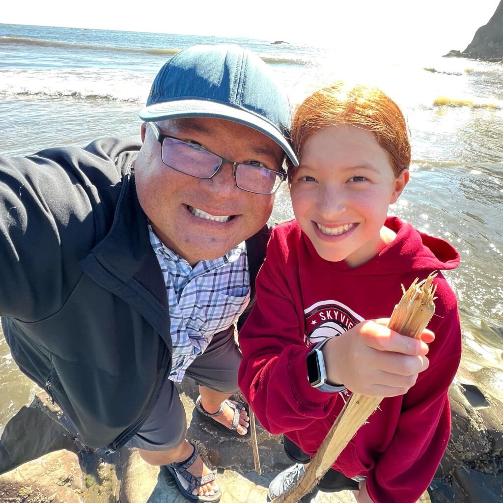 Two people smiling for a selfie on a rocky coastline with a person holding a piece of driftwood.