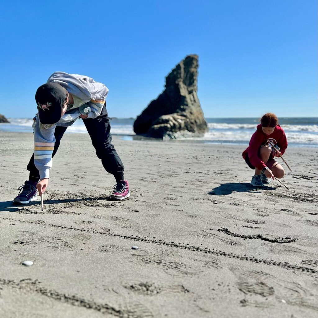 Two kids drawing in the sand on a sunny day at Cannon Beach, with the iconic Cannon Rock formation in the background. The ocean stretches out to the horizon, with waves gently breaking against the shore.