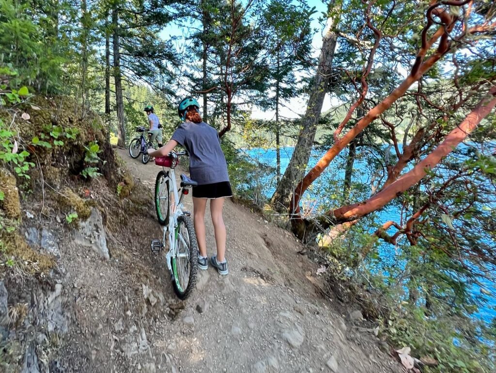 A cyclist pauses on a rocky trail overlooking a deep blue lake surrounded by tall evergreen trees.