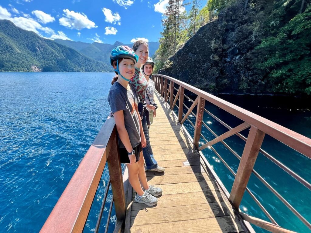 A family stands on a wooden suspension bridge with railings over a vibrant blue lake surrounded by mountains. The clear sky and lush greenery highlight a beautiful outdoor adventure.