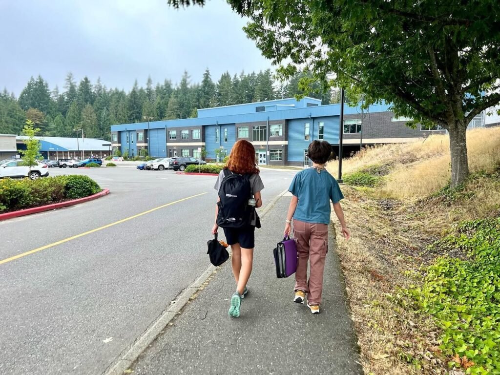 Two people walking away from a blue building with a backdrop of tall evergreen trees. A sidewalk and parking lot are visible on the left, with greenery in the foreground.