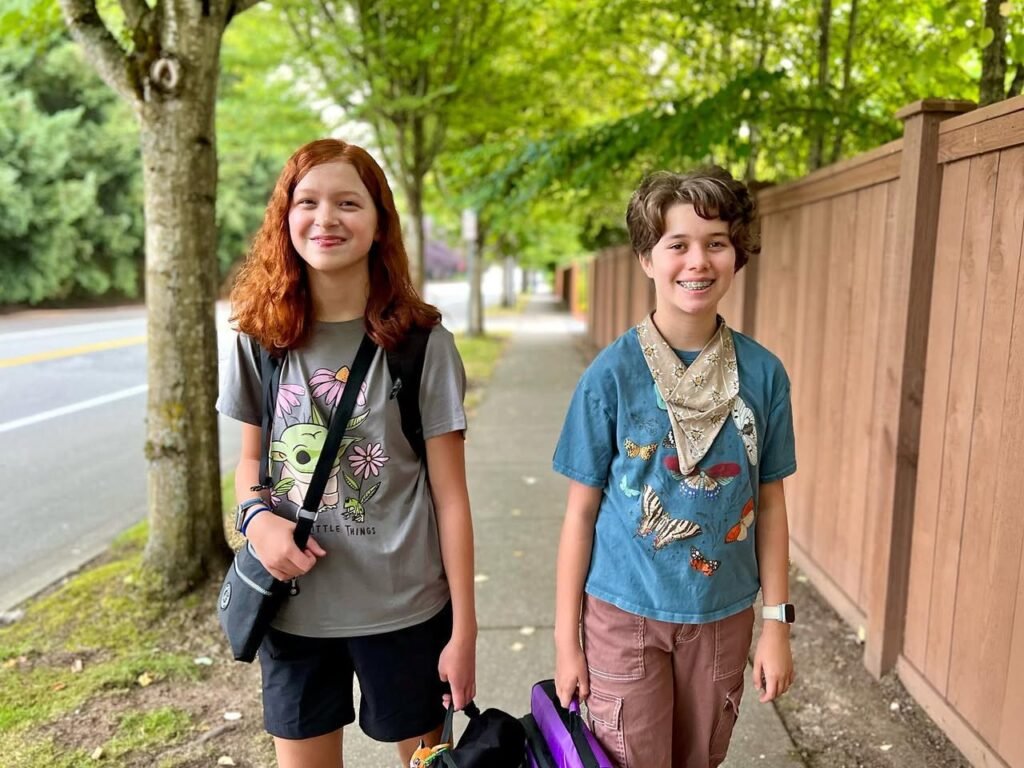 Two individuals smiling on a tree-lined sidewalk near a wooden fence. A close-up of a person standing by a tree in the background.