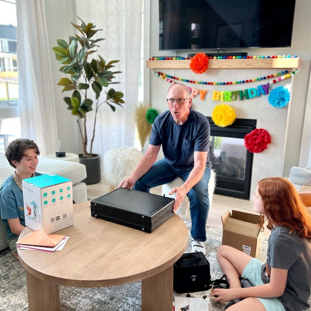 A man presents a birthday gift in a living room while two others watch. A birthday banner is displayed on the fireplace.