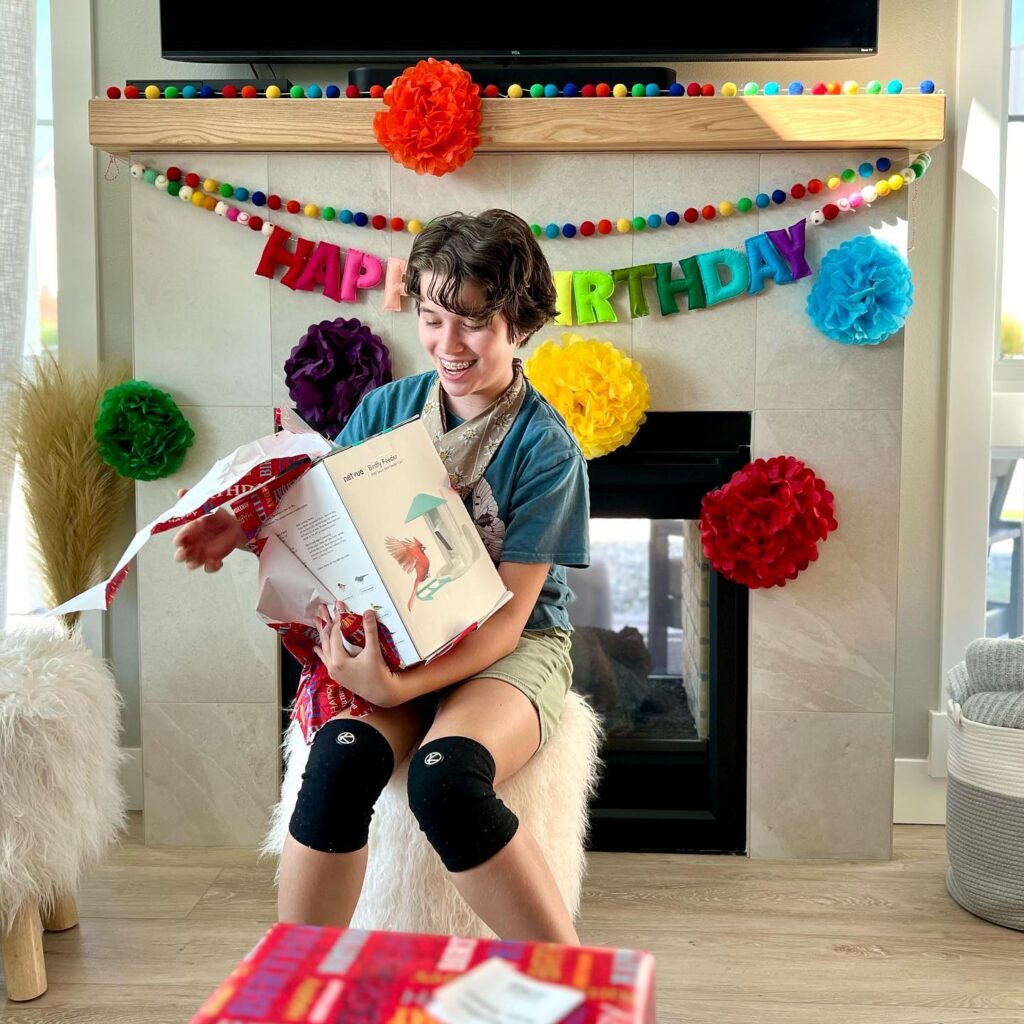 A person smiles while opening a book in a room decorated with a colorful Happy Birthday banner and paper pom-poms.