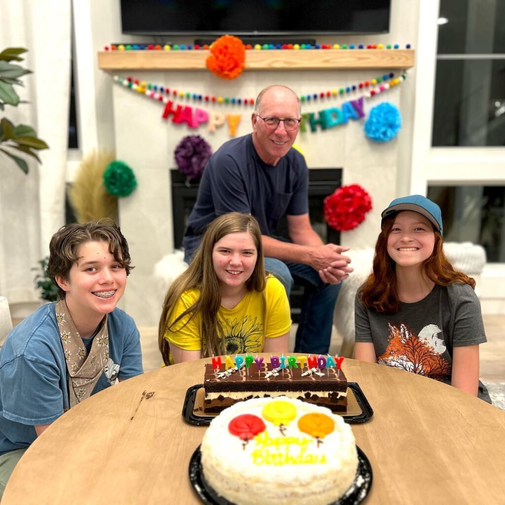 A family gathers around a table with two birthday cakes, celebrating a special day in a warmly decorated living room with a 'Happy Birthday' garland.