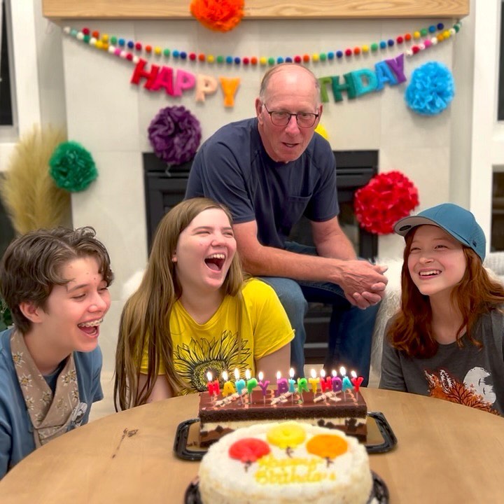 A group of family members celebrates a birthday with two cakes and a "Happy Birthday" banner at a table and by a fireplace.