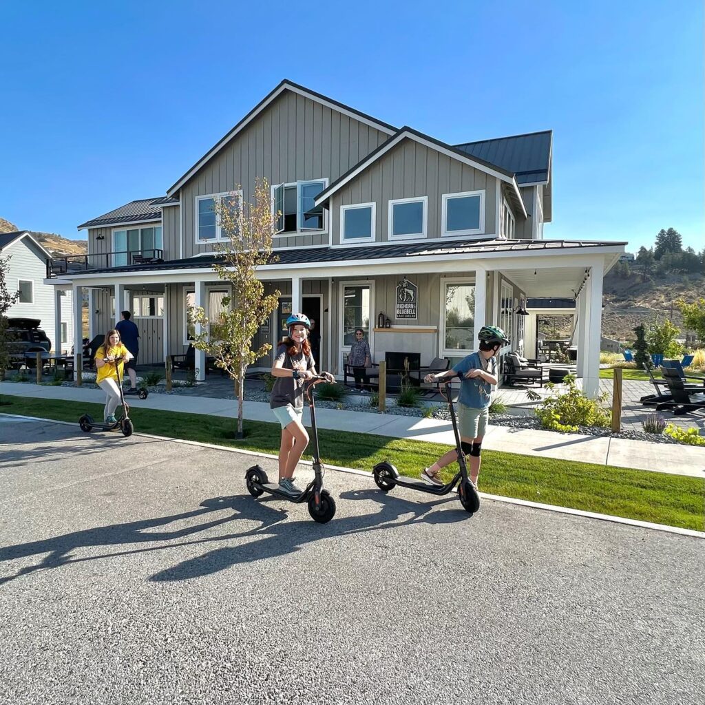 People enjoying a sunny day riding electric scooters on a suburban driveway outside a modern home.