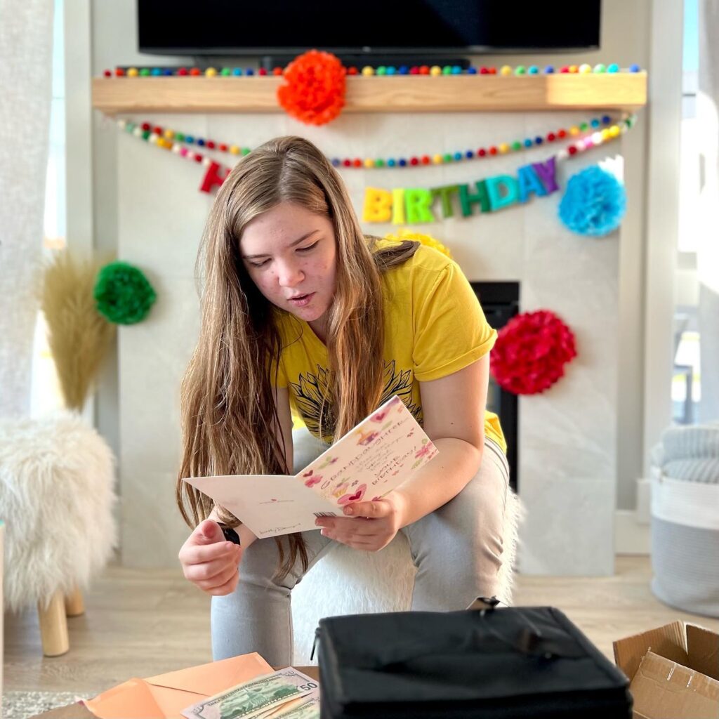 A person in a yellow shirt is kneeling on the floor and reading a hand-written birthday card. The background is decorated with a colorful banner and pom-poms.