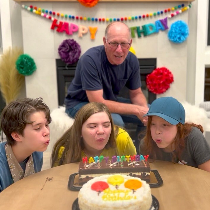A family gathered around a birthday cake with candles, ready to blow them out on a day filled with smiles and laughter, decorated with colorful bunting and pom-poms.