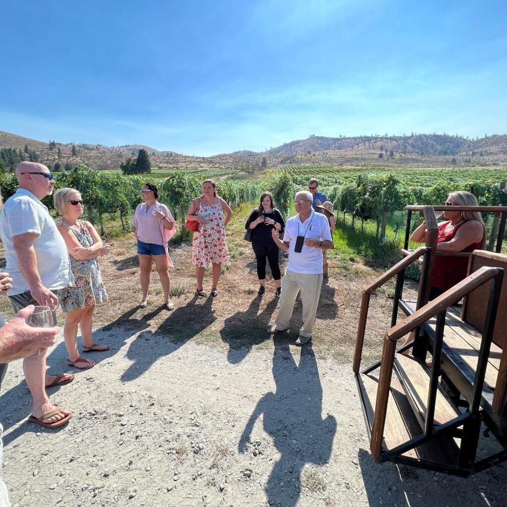 A group of people stand outside enjoying a wine tasting at a vineyard. Lush green vines stretch out under a bright blue sky.
