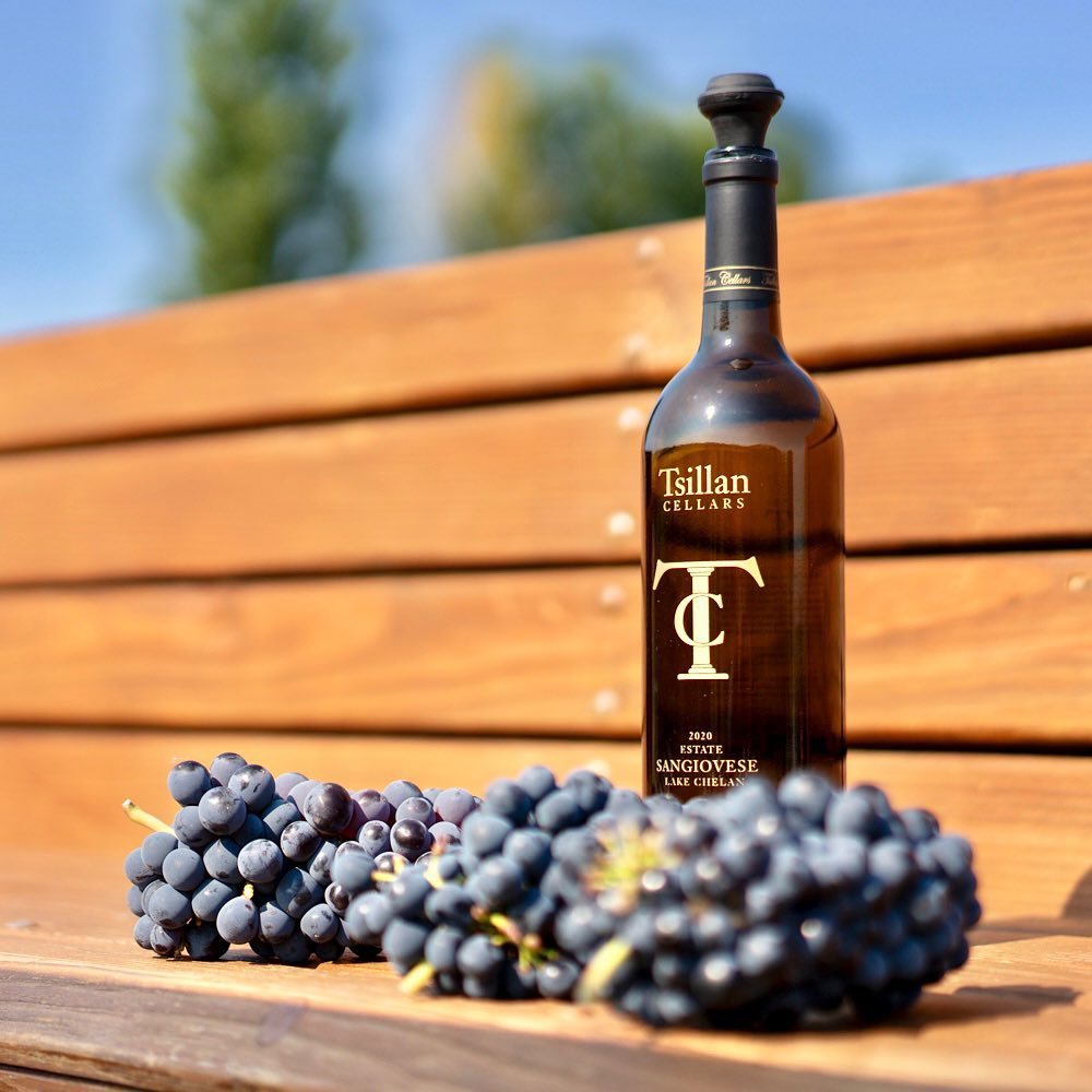 A bottle of Tsillan winery pinot noir wine beside fresh grape clusters, placed on a wooden bench outdoors.