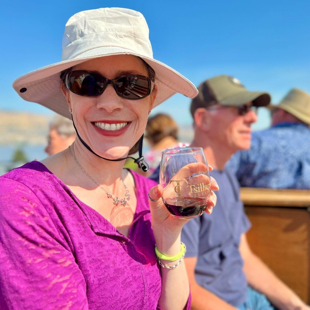 A woman in a hat and sunglasses holds a glass of red wine outdoors under a clear blue sky.