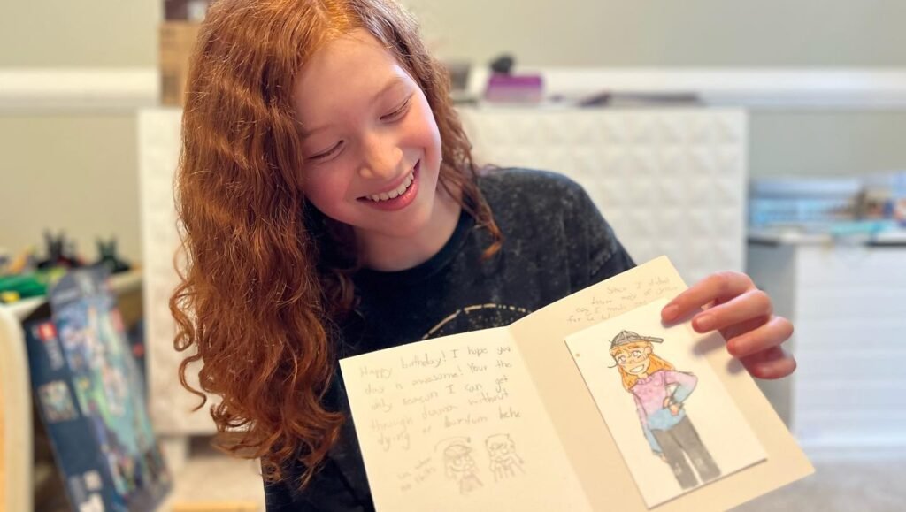 A person with curly red hair smiles while looking at a handmade book containing a colorful drawing.