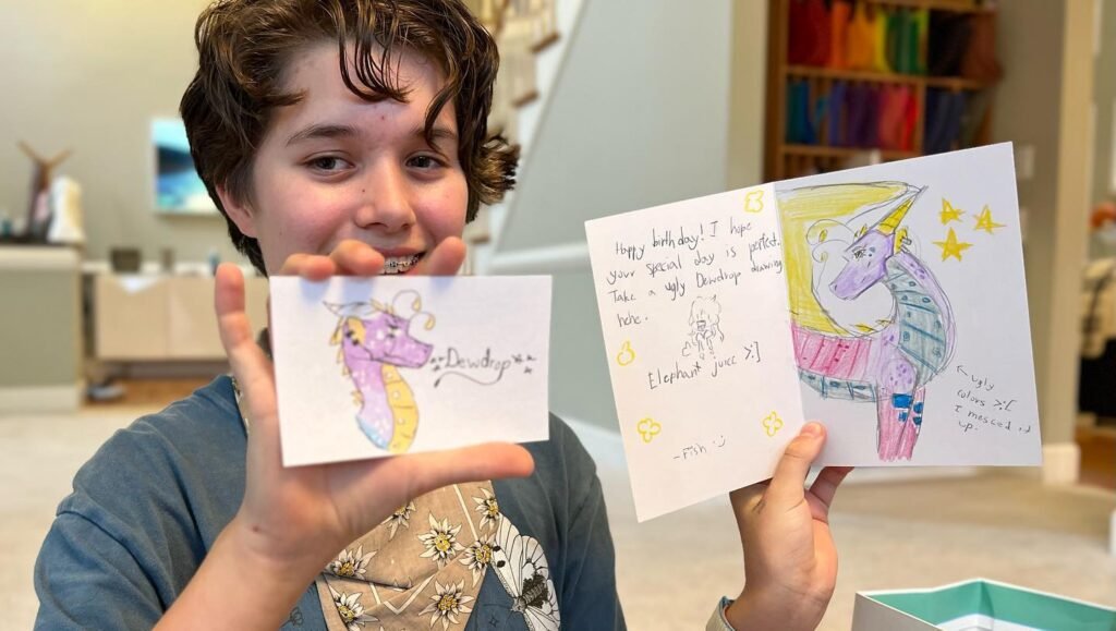 A child proudly holding up hand-drawn cards featuring colorful unicorn illustrations and a written message.