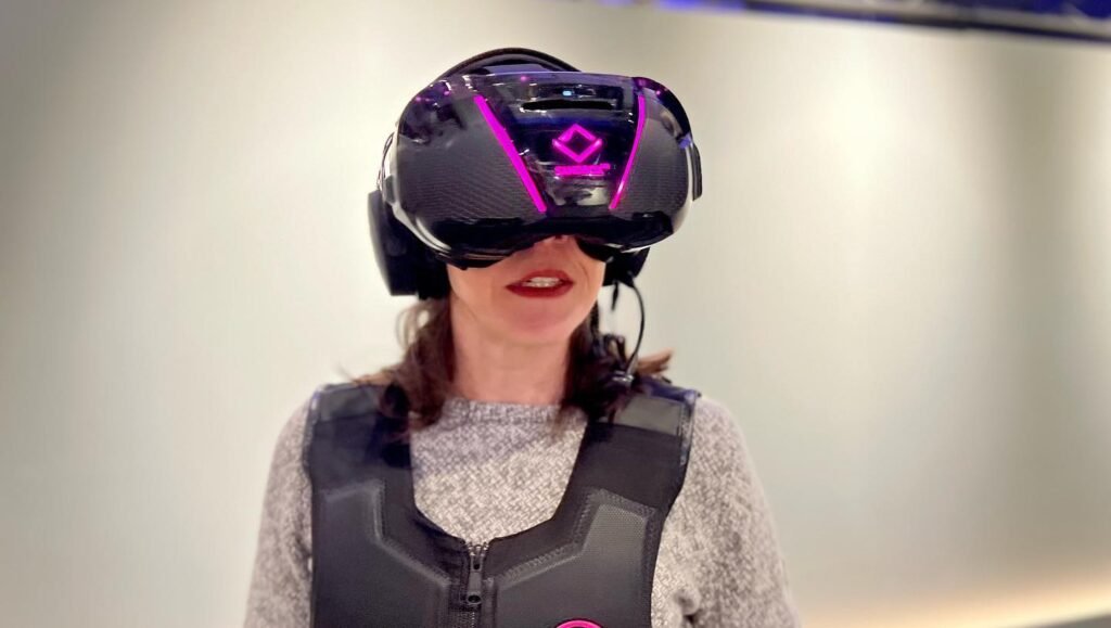 A person wearing a VR headset and haptic vest, smiling during a virtual reality gaming session. The setup includes a detailed VR visor and specialized gear.