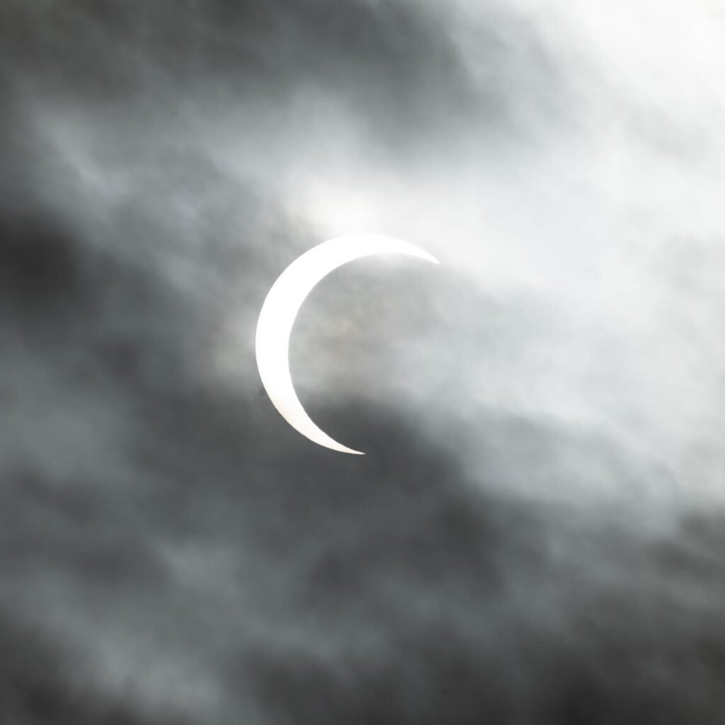 The moon is positioned in front of the sun, partially obscuring its face and creating a crescent shape. The scene is framed by a variety of gray and white clouds.