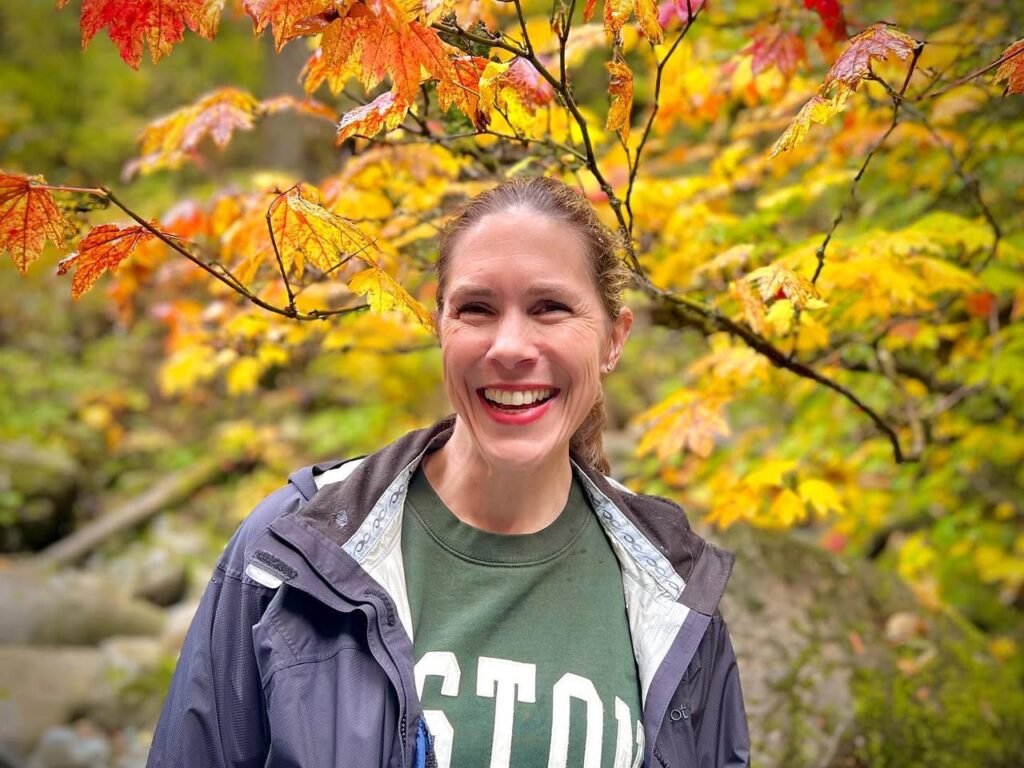 A person smiles in front of vibrant fall foliage. The scene includes a stone wall or landscape element.