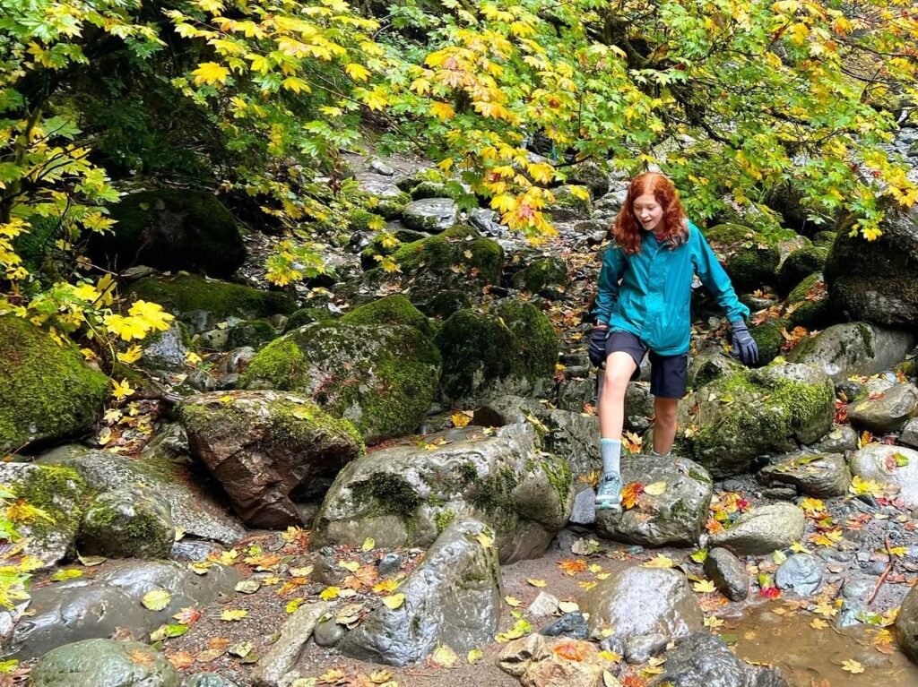 A hiker navigates a rocky stream bed surrounded by vibrant autumn foliage and moss-covered rocks.