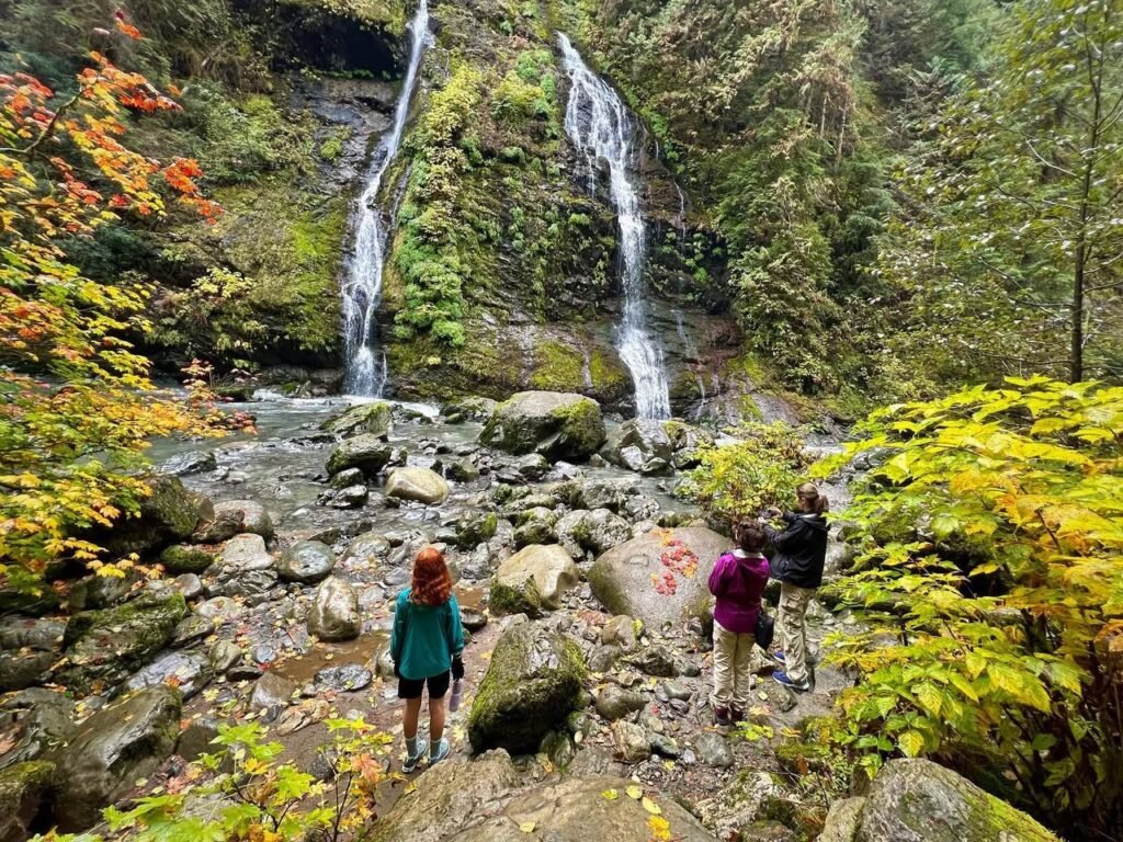 Hikers walking among rocks towards a split waterfall with autumn foliage around the forested area.