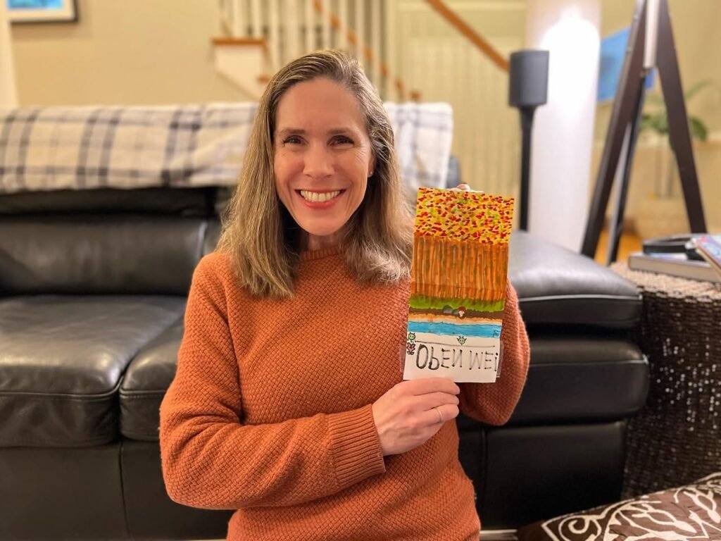 A woman proudly displays a handmade race ribbon in an orange sweater.