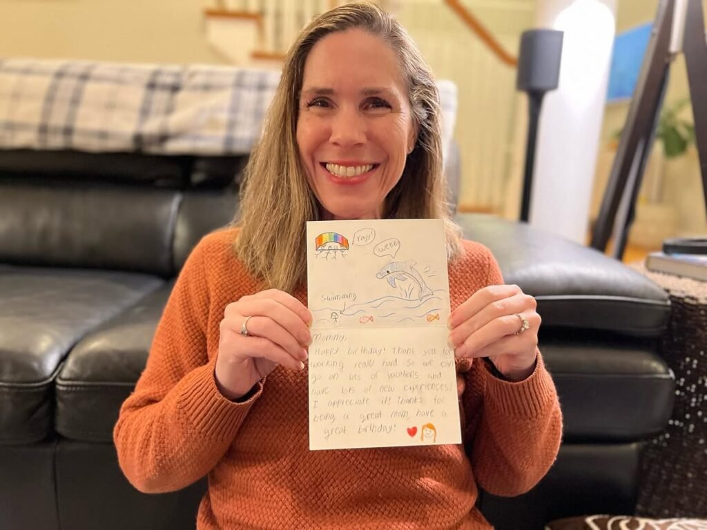 A smiling woman in an orange sweater holds up a handwritten card in her cozy living room.