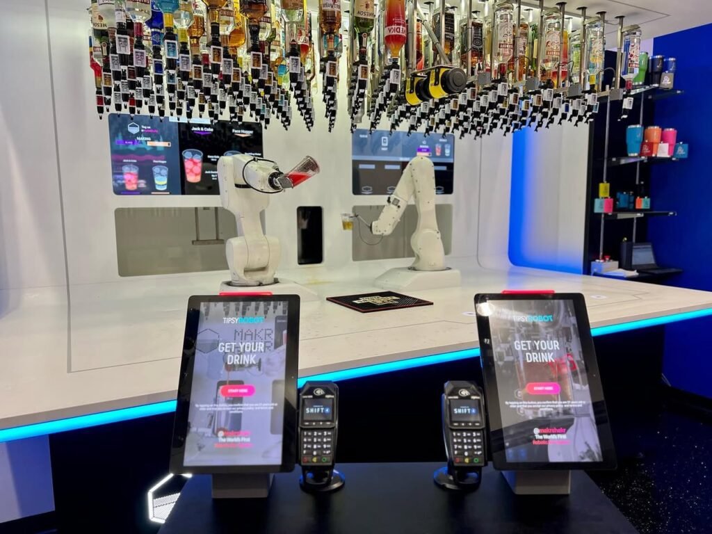 A futuristic bar counter featuring white robotic arms preparing beverages with overhead drink modules and digital payment tablets.