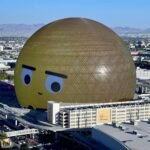 An aerial view of the large spherical structure known as The Sphere in Las Vegas. The building features a golden exterior and a face printed on its surface.