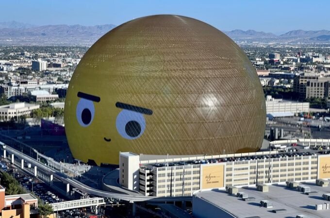 An aerial view of the large spherical structure known as The Sphere in Las Vegas. The building features a golden exterior and a face printed on its surface.