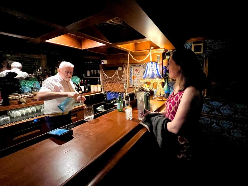 A customer sits at a dark wooden bar counter while a bartender prepares a drink. The bar is warmly lit with decorative blue lamps in the background.