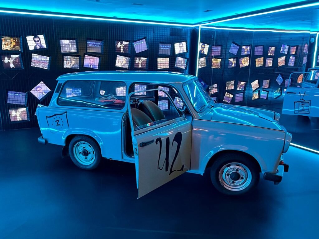 A vintage car with the number 212 on its door is displayed under blue neon lighting with a wall of framed photos.