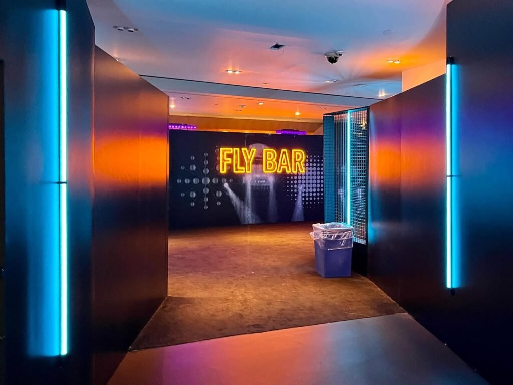 The Fly Bar features bold blue lighting and a glowing neon sign against warm orange light. Accessible through a modern, stylish interior corridor.