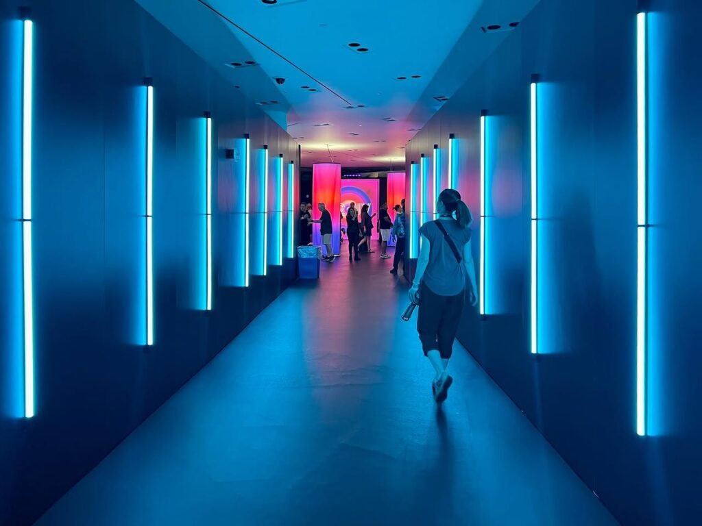 A person walks through a futuristic corridor with vertical blue wall lights. Pink and orange light emanates from the far end of the hallway.
