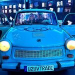 Two people sit inside a blue Trabant with U2 branding, lit by blue lights.