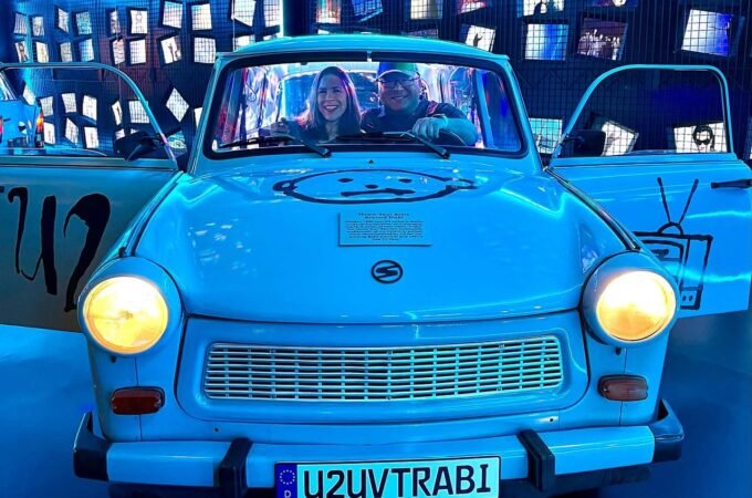 Two people sit inside a blue Trabant with U2 branding, lit by blue lights.