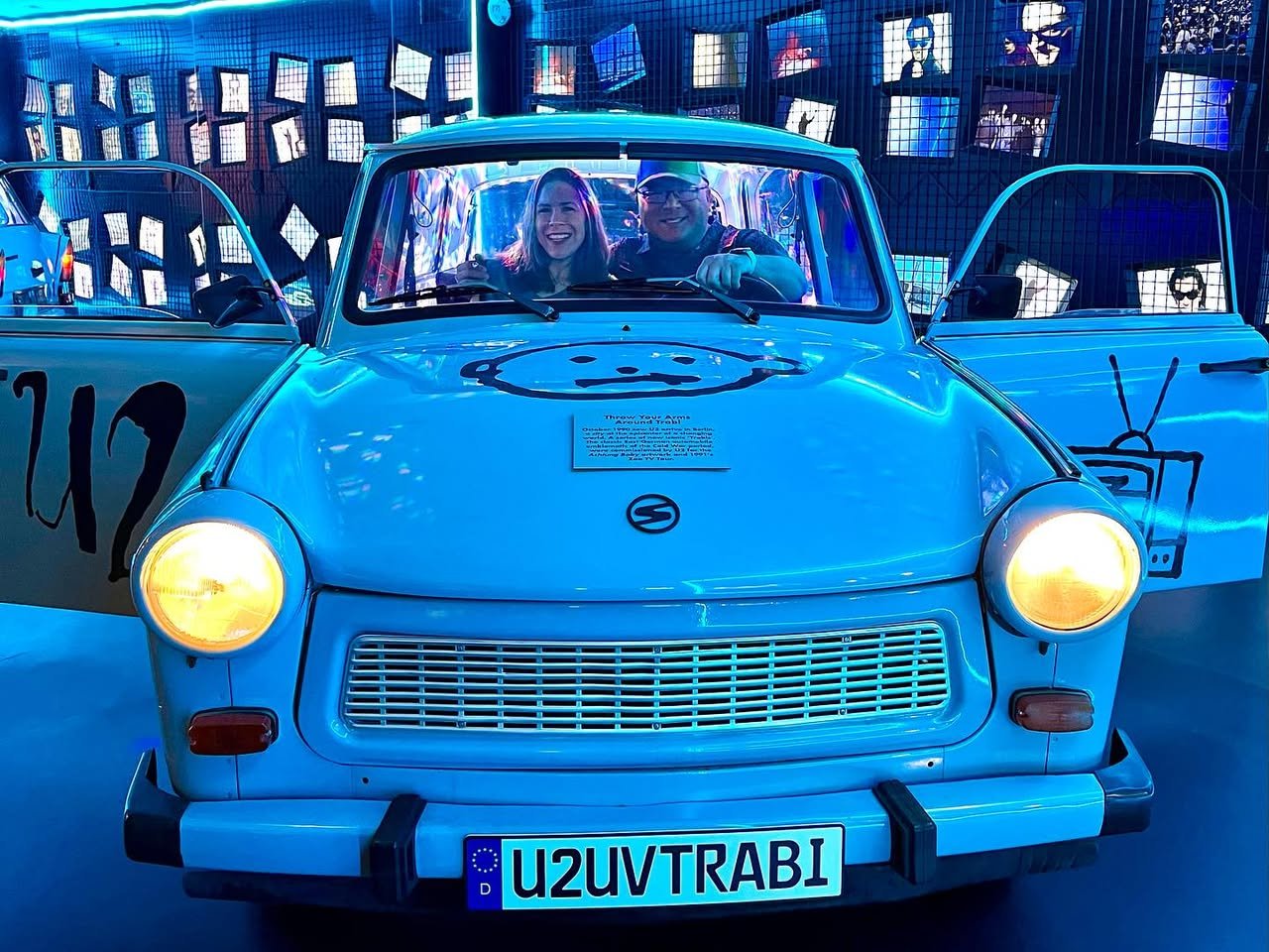 U2 Zoo Station Exhibit Two people sit inside a blue Trabant with U2 branding, lit by blue lights.