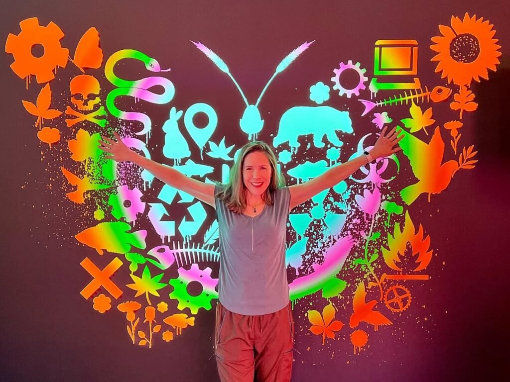 A person stands in front of a vibrant butterfly-shaped art display featuring nature and tech symbols. Textured close-up shows neon hangers and colors.