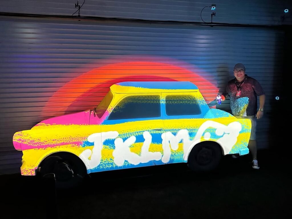 Person using a UV light projector to create a vibrant, neon-colored car design on a metal wall at night.