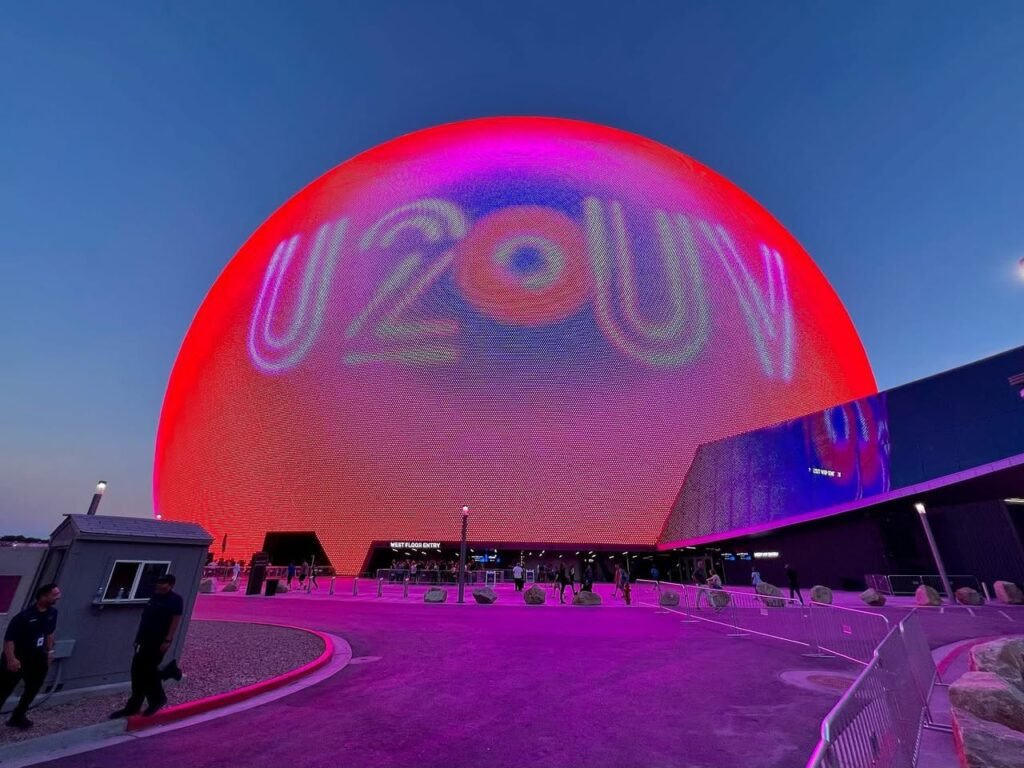 The Space Needle in Seattle is illuminated in red and purple with the word 'UFO' projected on its sides. Visitors walk along pathways beneath the glowing structure at twilight.