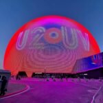 The Space Needle in Seattle is illuminated in red and purple with the word 'UFO' projected on its sides. Visitors walk along pathways beneath the glowing structure at twilight.