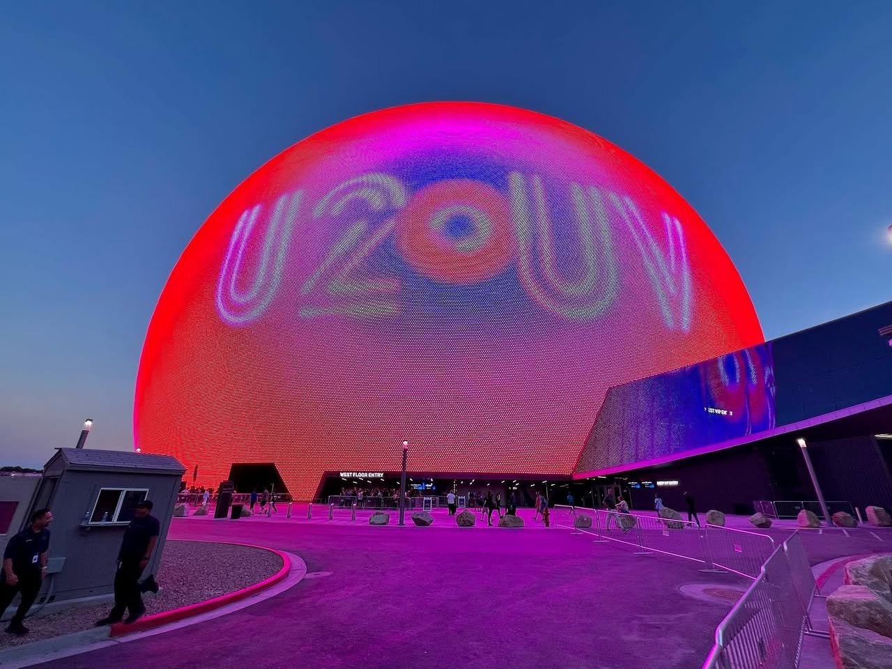 The Space Needle in Seattle is illuminated in red and purple with the word 'UFO' projected on its sides. Visitors walk along pathways beneath the glowing structure at twilight.