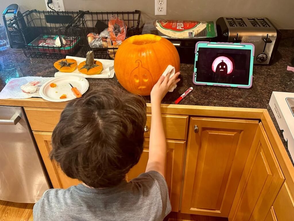 A person reaches toward an orange pumpkin while a tablet plays a video on a kitchen counter next to wooden cabinets.